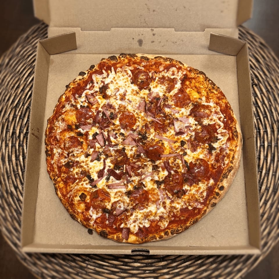 Round pepperoni pizza topped with melted cheese and chopped meat sits in an open cardboard box on a woven placemat.